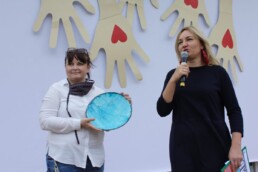 Zdjęcie, dwie kobiety na scenie; jedna mówi przez mikrofon, druga trzyma ozdobny talerz