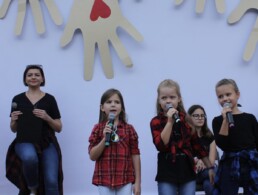 Zdjęcie, trzy śpiewające dziewczynki w trakcie występu, w tle reszta zespołu