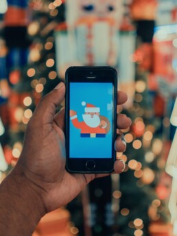Zdjęcie; dłoń trzymająca smartfon; na ekranie zdjęcia obrazek Mikołaja; w tle świąteczne światełka