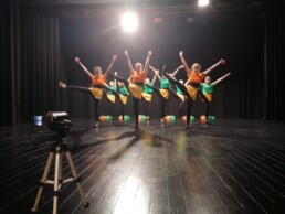 Zdjęcie_Grupa taneczna młodych dziewczyn tańczy na scenie. Prawe nogi mają podniesione w wysoko i ręce są wyciągnięte do góry. Kamera na statywie stoi w lewym dolnym rogu.
