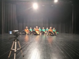 Zdjęcie_ Młode dziewczyny tańczą na scenie, siedzą i mają przełożoną lewą nogę. Na pierwszym planie po lewej stoi statyw z kamerą.