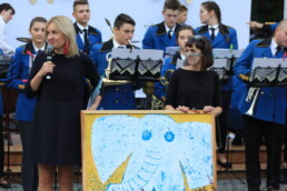Zdjęcie, dwie kobiety stoją między obrazem, na którym namalowany jest słoń; w tle - orkiestra dęta