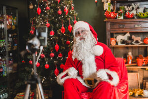 Zdjęcie, święty Mikołaj ubrany w czerwony strój, siedzi na fotelu; w tle choinka z czerwonymi ozdobami i regał ze świątecznymi upominkami