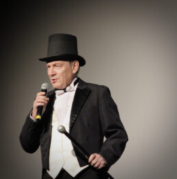 Zdjęcie_ Starszy mężczyzna, ubrany w smoking i kapelusz, stoi na scenie i przemawia do mikrofonu. W drugiej ręce trzyma metalowy element. Zbliżenie od pasa w górę.