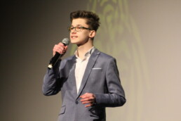 Zdjęcie_ Młodzieniec w okularach, ubrany w garnitur stoi na scenie i przemawia do mikrofonu. Zbliżenie od pasa w górę.