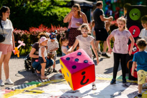 Dzieci na zewnątrz bawią się na kolorowej planszy rozłożonej na ziemi, na pierwszym planie dziewczynka trzyma kolorową kostkę 