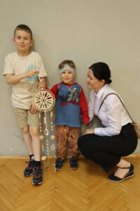 Na zdjęciu widocznych jest 2 chłopczyków i pani, jeden z chłopczyków trzyma w ręku zrobiony z drewnianej sklejki dzwonek wietrzny  